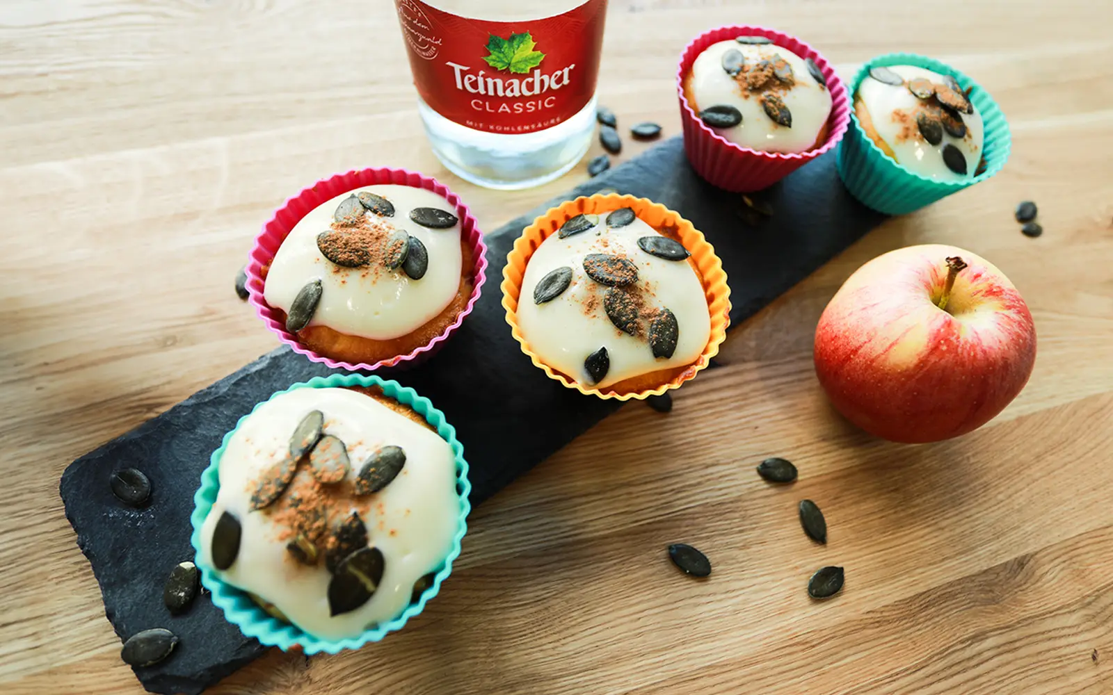 Kürbis-Muffins mit cremigem Zuckerguss, Kürbiskernen und Zimt. Serviert mit einem Apfel und Teinacher Classic