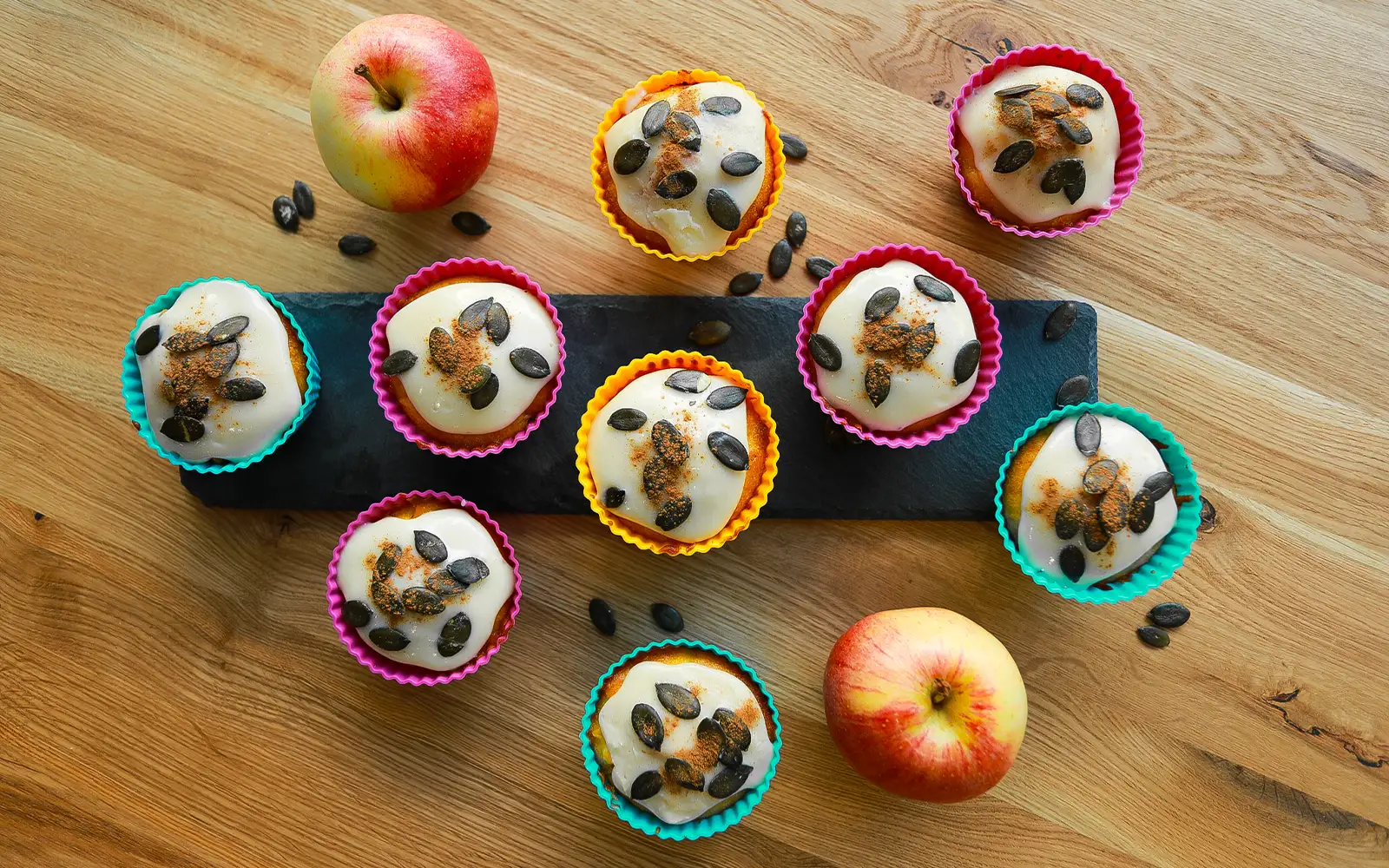 Saftige Kürbis-Muffins mit Zuckerguss, Kürbiskernen und Zimt, serviert in bunten Förmchen auf einer Schieferplatte