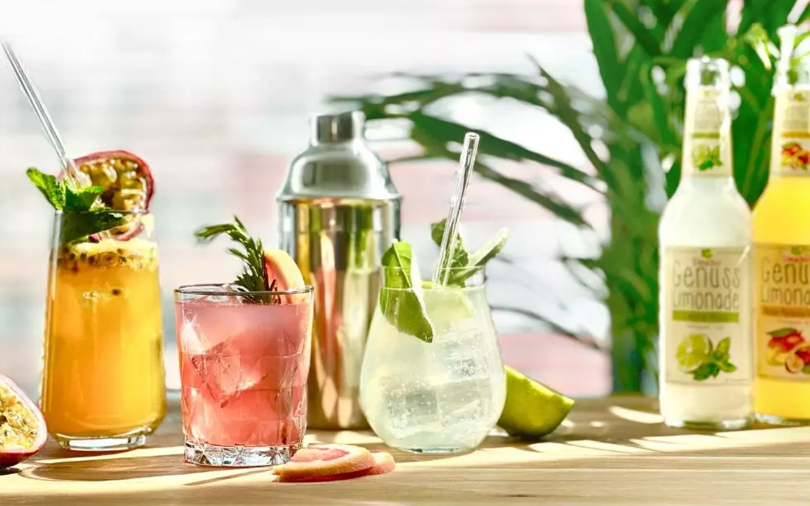 teinacher-blog-mocktail-magie-teinacher-rezepte Drei bunte Mocktails mit Teinacher Genuss-Limonade, frischen Früchten und Kräutern auf einem Holztisch neben einem Shaker.
