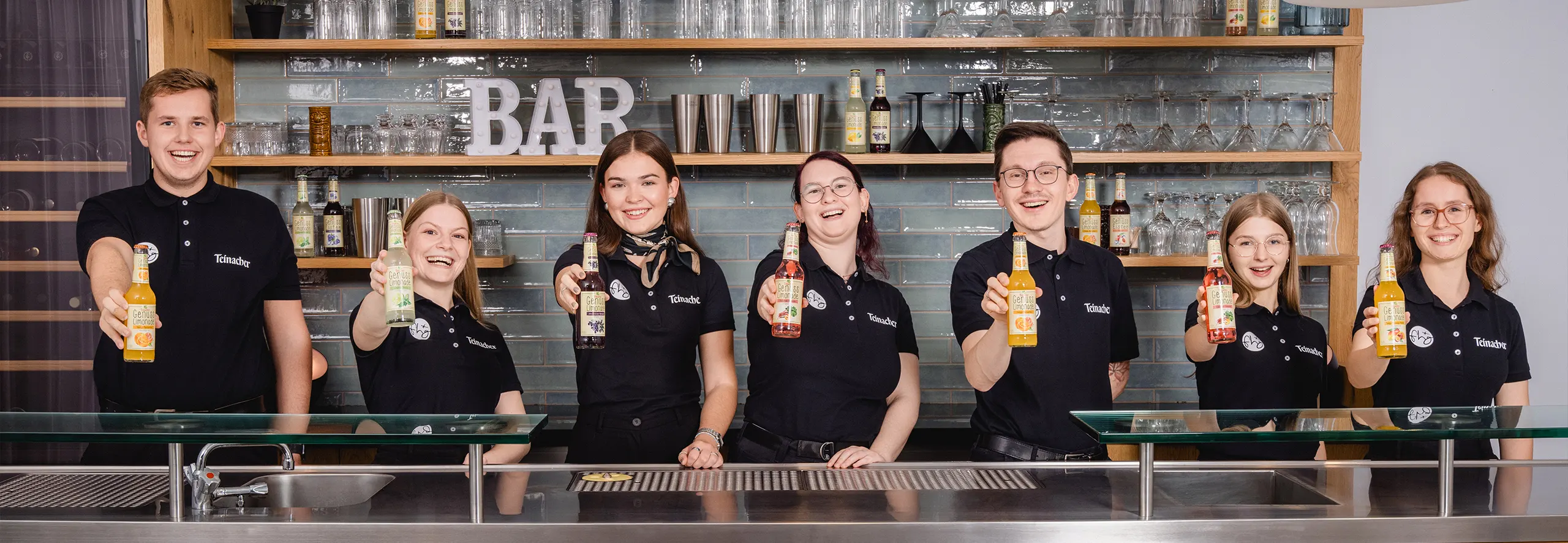 Auszubildende von Teinacher und der FHG stehen hinter einer Bar und halten verschiedene Teinacher Genuss-Limonaden in die Kamera.