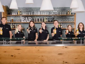 Auszubildende von Teinacher und der FHG stehen hinter einer Bar und halten verschiedene Teinacher Genuss-Limonaden in die Kamera.