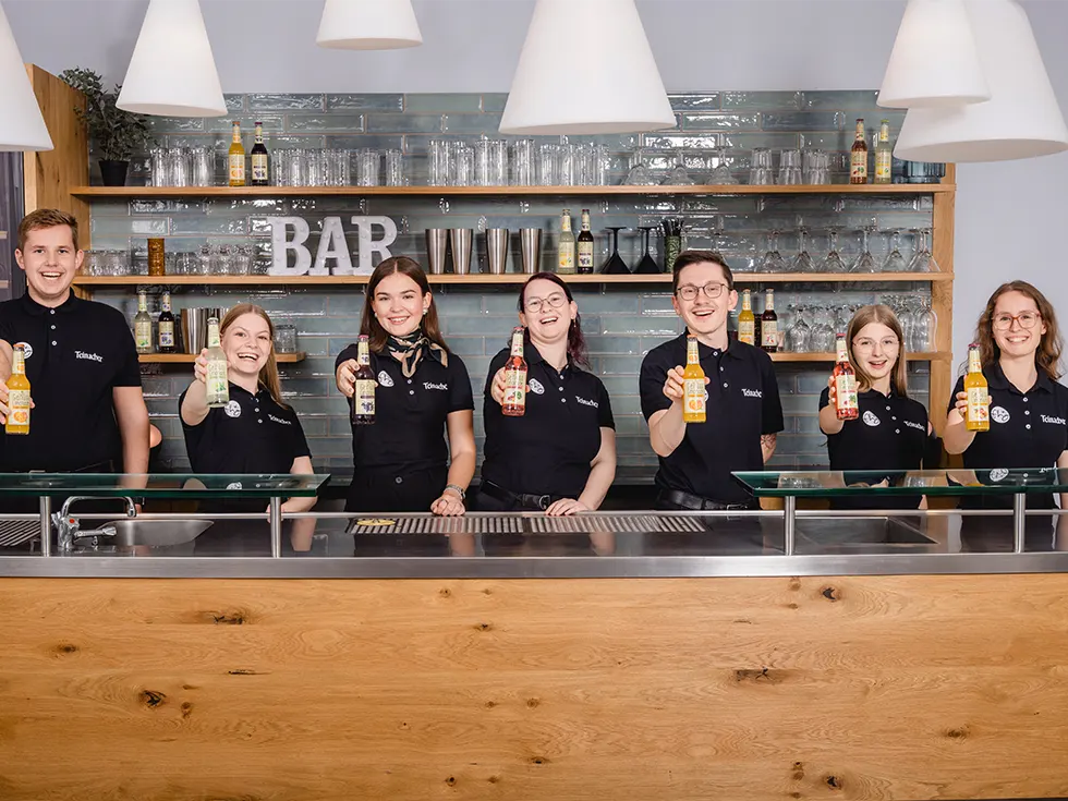 Auszubildende von Teinacher und der FHG stehen hinter einer Bar und halten verschiedene Teinacher Genuss-Limonaden in die Kamera.