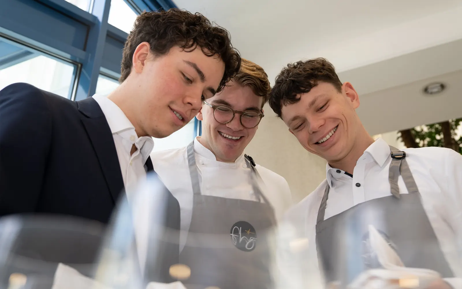 Drei Auszubildende der FHG stehen gemeinsam in Arbeitskleidung und schauen lächelnd auf eine Tätigkeit im gastronomischen Umfeld.