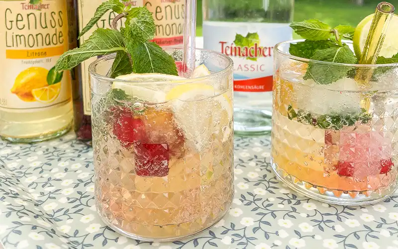 Mocktail in 2 Gläsern und Früchte mit Genuss-Limonaden im Hintergrund