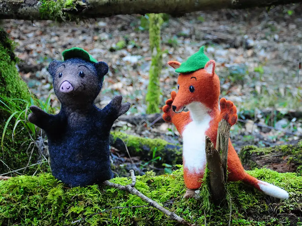Zwei Filzfiguren, ein Bär und ein Fuchs mit grünen Hüten, stehen in einem Wald auf Moos