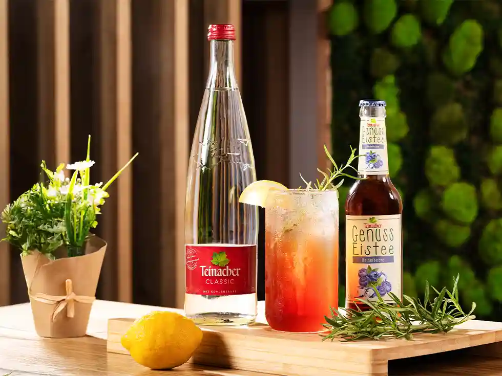 Teinacher Classic, Teinacher Genuss-Eistee Heidelbeere und ein erfrischender Eistee-Cocktail mit Zitrone und Rosmarin auf einem Holzbrett, dekoriert mit Kräutern und einer Zitrone.