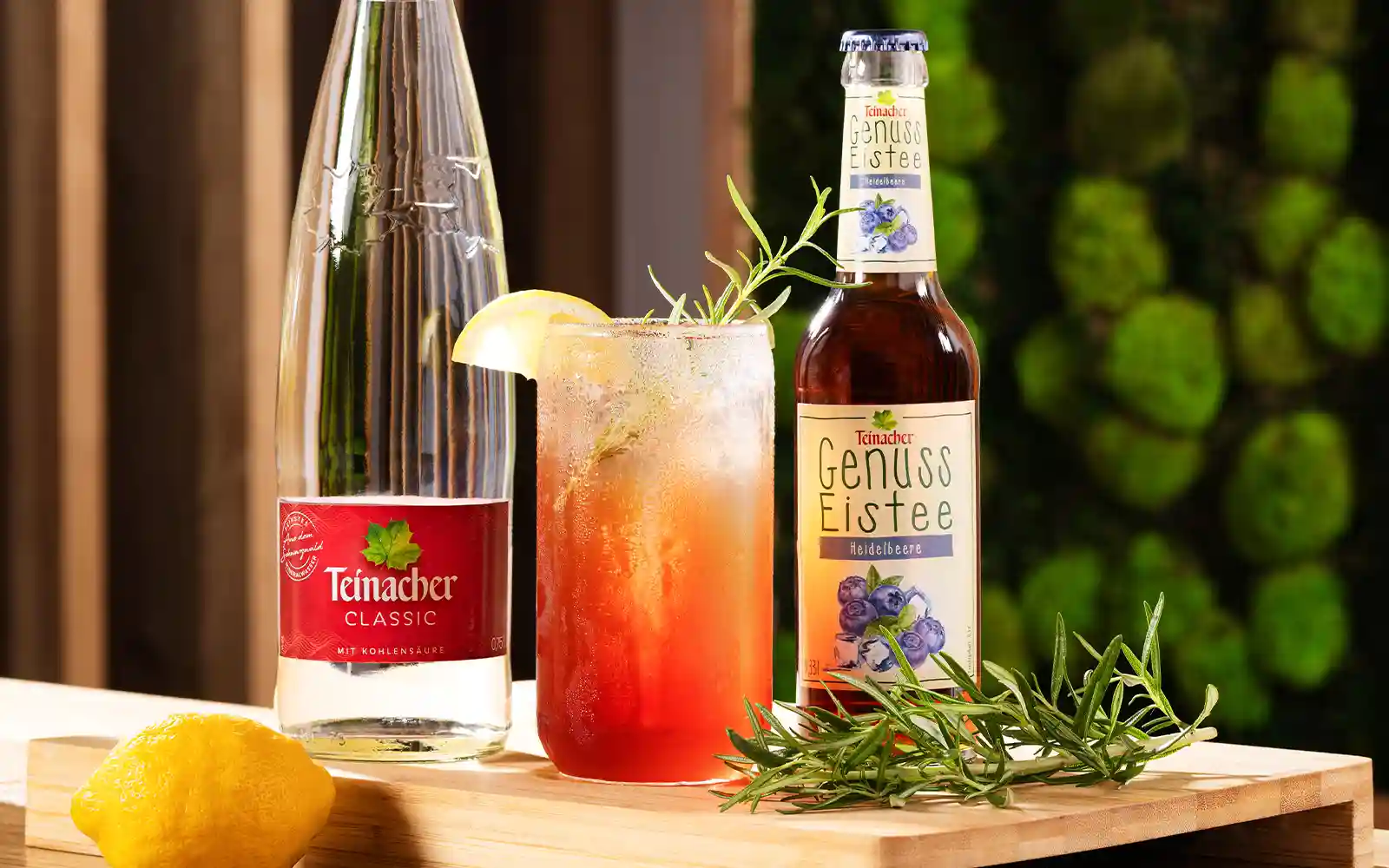 Teinacher Classic, Teinacher Genuss-Eistee Heidelbeere und ein erfrischender Eistee-Cocktail mit Zitrone und Rosmarin auf einem Holzbrett, dekoriert mit Kräutern und einer Zitrone.