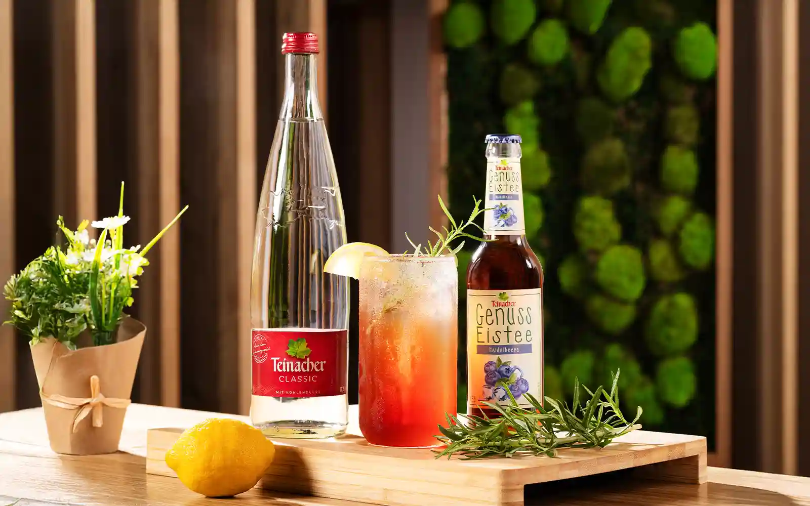Teinacher Classic, Teinacher Genuss-Eistee Heidelbeere und ein erfrischender Eistee-Cocktail mit Zitrone und Rosmarin auf einem Holzbrett, dekoriert mit Kräutern und einer Zitrone.