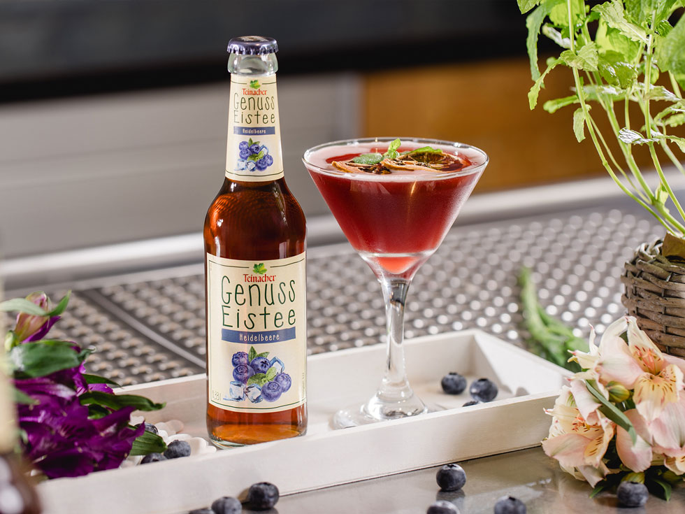Flasche Teinacher Genuss Eistee Heidelbeere neben einem roten Cocktailglas auf einem Tablett, umgeben von Heidelbeeren und Blumen