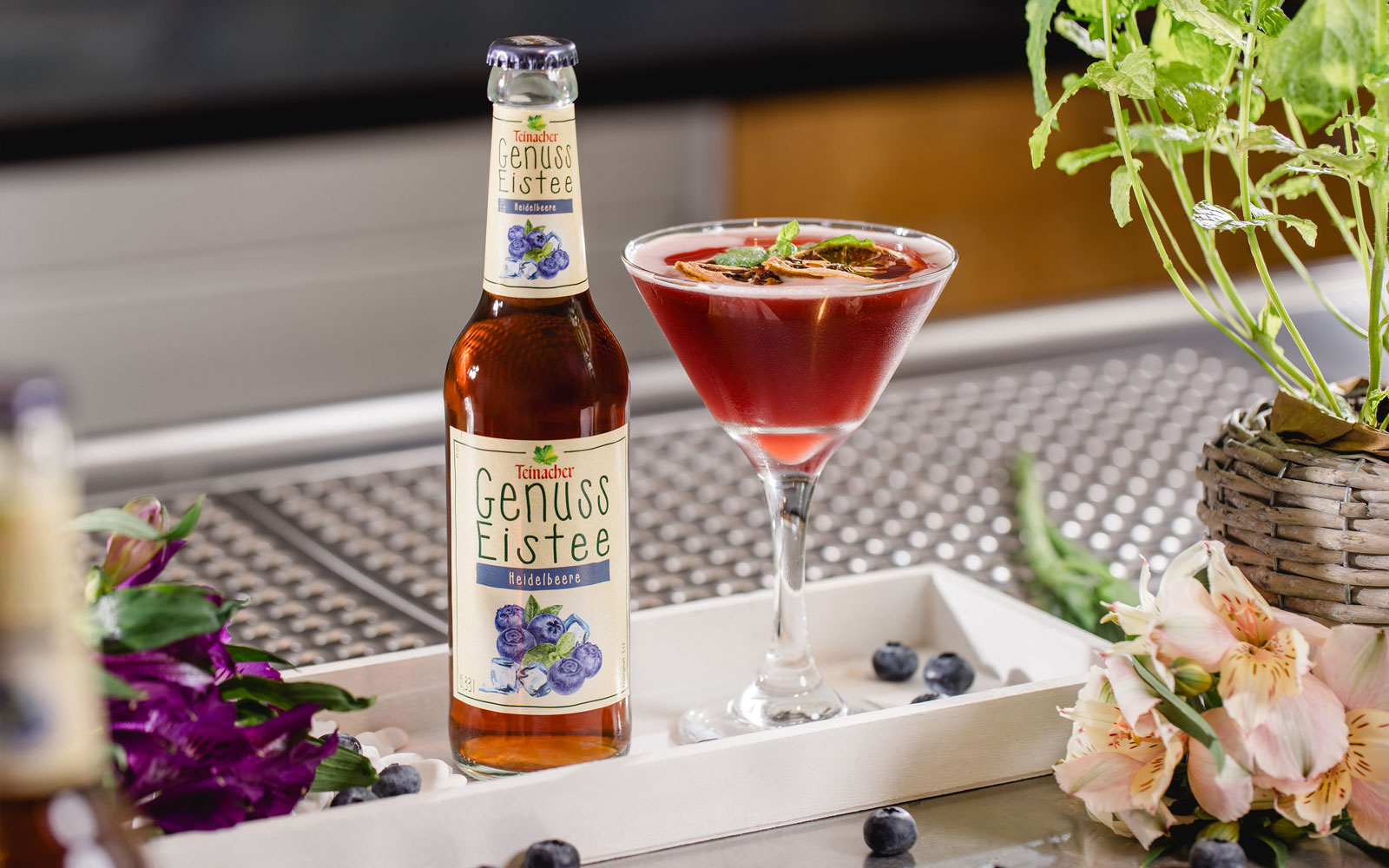 Flasche Teinacher Genuss Eistee Heidelbeere neben einem roten Cocktailglas auf einem Tablett, umgeben von Heidelbeeren und Blumen