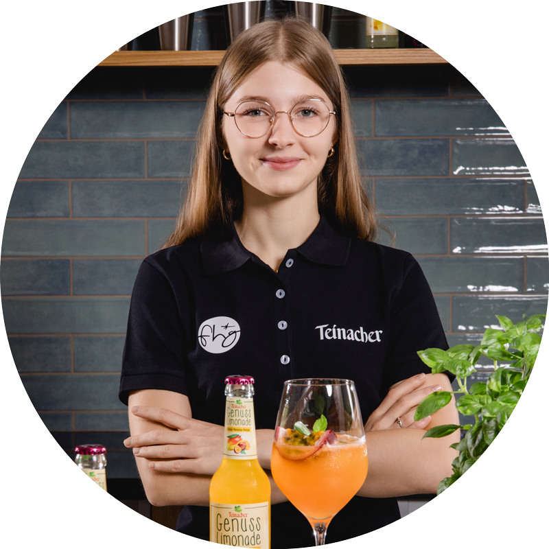 Frau mit Brille und verschränkten Armen steht hinter einer Theke mit Teinacher Getränkeflasche und gefülltem Glas, im Hintergrund Regal und Fliesenwand