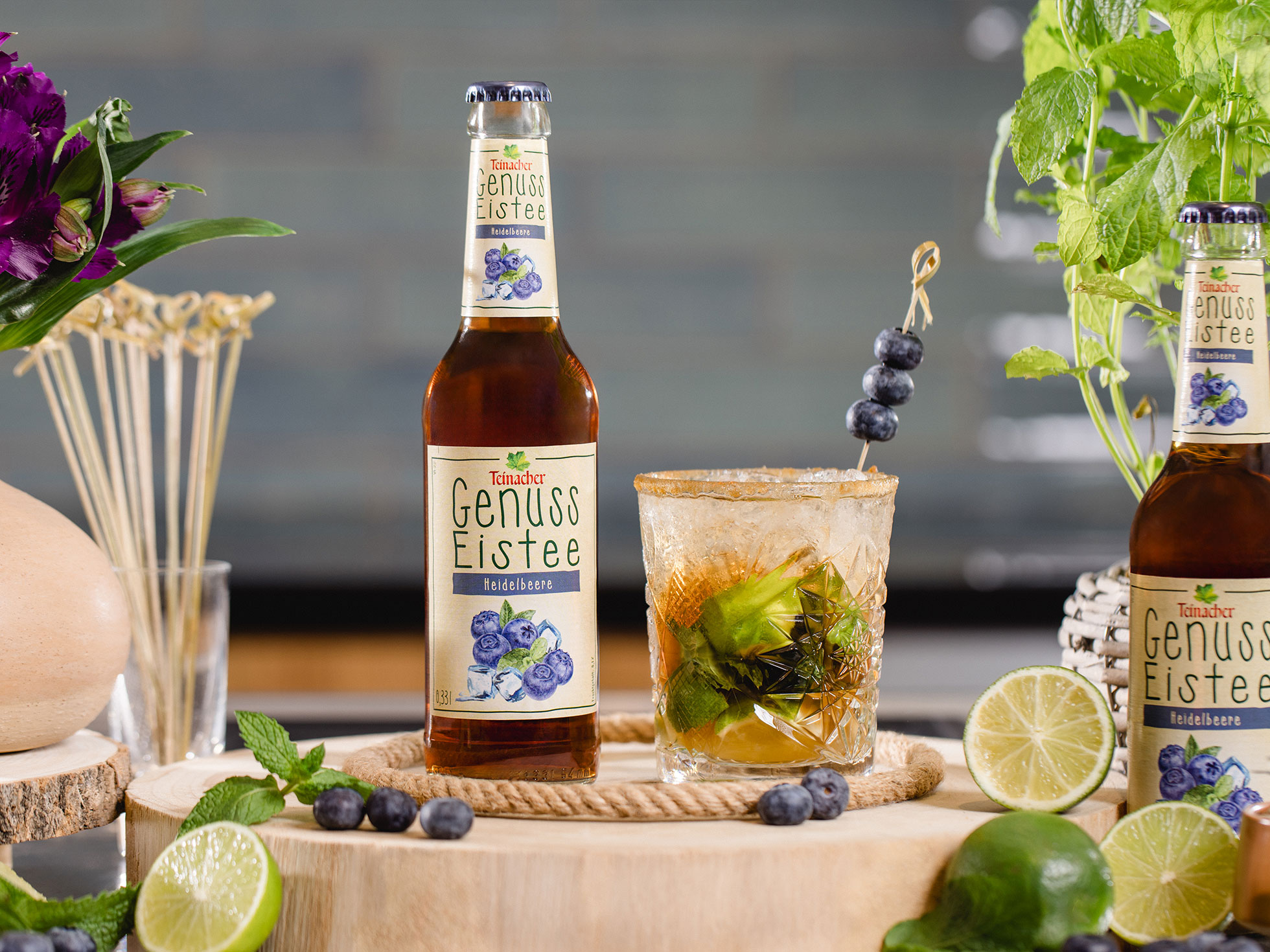Flasche Teinacher Genuss Eistee Heidelbeere neben einem Glas mit Eis, Limette, Minze und Heidelbeeren auf einer Holzscheibe