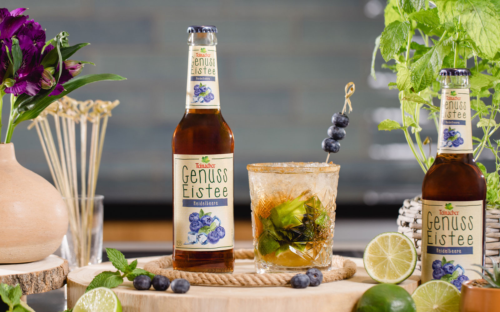 Flasche Teinacher Genuss Eistee Heidelbeere neben einem Glas mit Eis, Limette, Minze und Heidelbeeren auf einer Holzscheibe