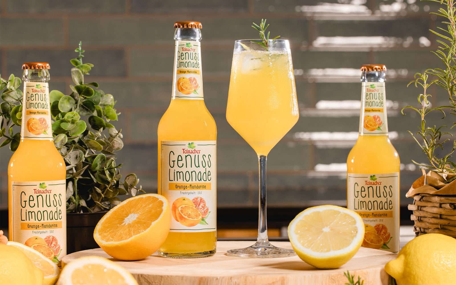 Zwei Flaschen Teinacher Genuss-Limonade Orange-Mandarine neben einem Glas mit Eis und Rosmarin, umgeben von Orangen- und Zitronenhälften.
