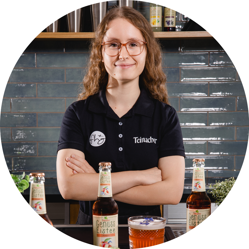 Frau mit Brille und verschränkten Armen steht hinter einer Theke mit Teinacher Getränkeflaschen und einem Glas, im Hintergrund Regale und Fliesenwand