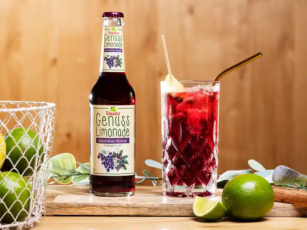 Teinacher Genusslimonade Schwarze Johannisbeere-Holunder mit Glas, Limetten und Trinkhalm.
