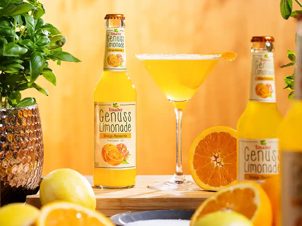 Teinacher Genusslimonade Orange-Mandarine mit Glas, frischen Orangen und Zitronen, dekorativ präsentiert.