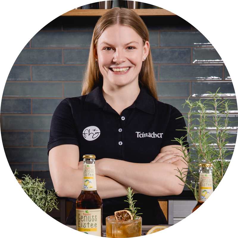 Frau mit verschränkten Armen steht hinter einer Theke mit Teinacher Getränkeflasche, Glas mit Eis und Rosmarin, im Hintergrund Regal und Fliesenwand