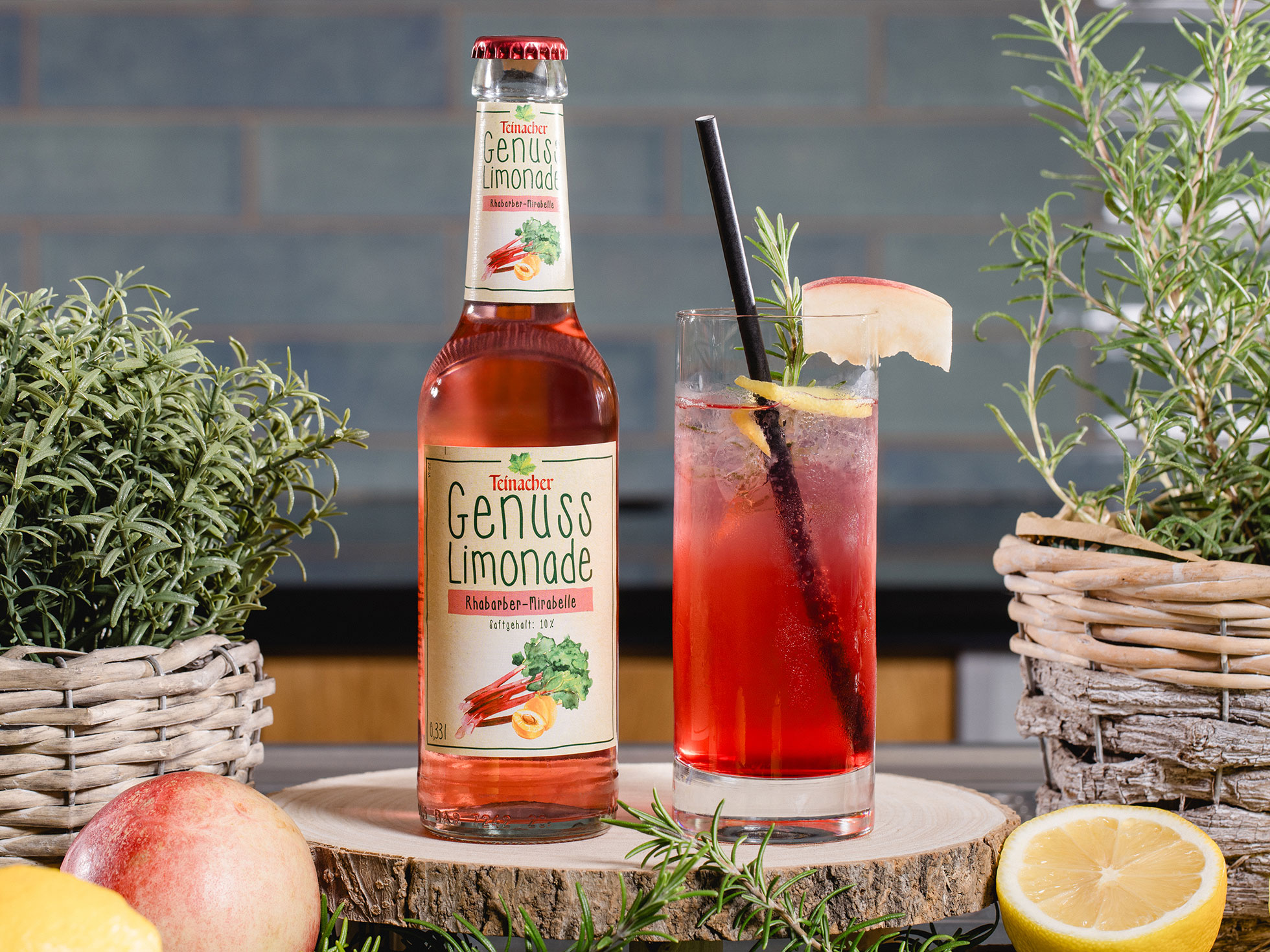 Flasche Teinacher Genuss Limonade Rhabarber-Mirabelle neben einem Glas mit Eis, Rosmarin und Fruchtgarnitur, umgeben von Kräutern und Zitrusfrüchten