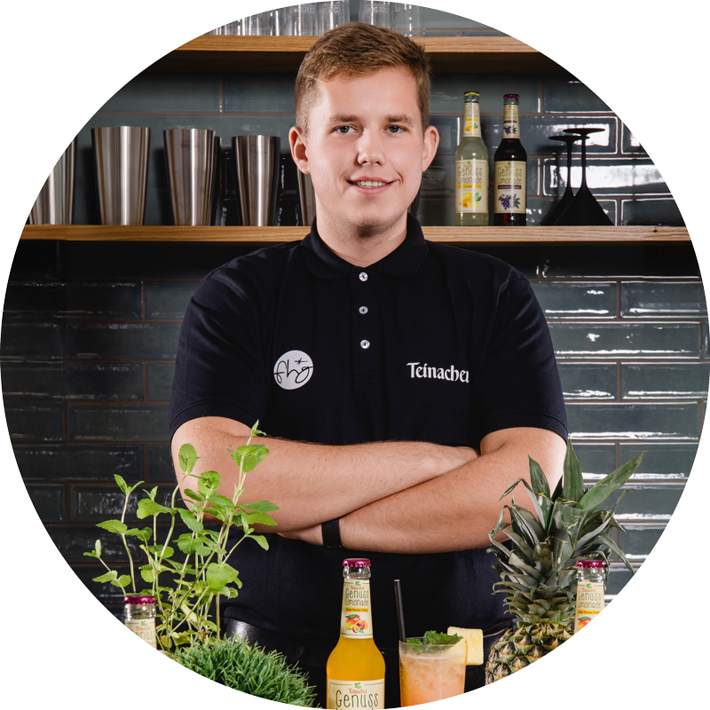 Mann mit verschränkten Armen steht hinter einer Theke mit Teinacher Getränkeflaschen, Kräutern und Früchten, im Hintergrund Regale und Fliesenwand