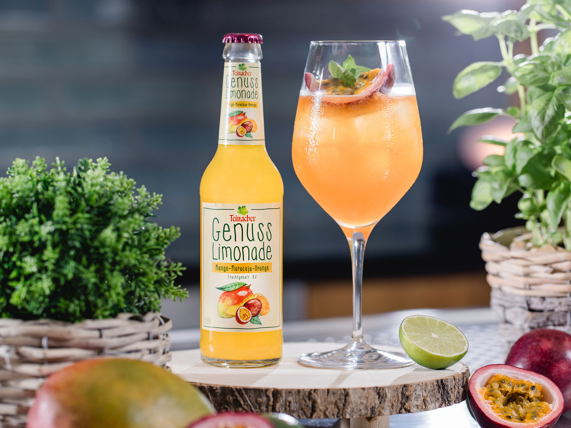 Flasche Teinacher Genuss Limonade Mango-Maracuja-Orange neben einem gefüllten Glas mit Eis, Minze und Maracuja, umgeben von Kräutern und Limette