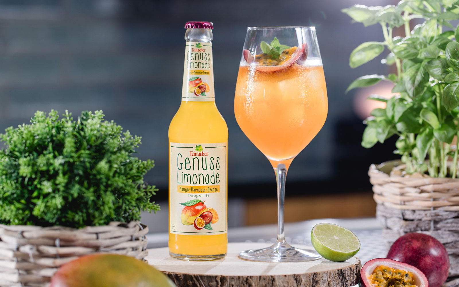 Flasche Teinacher Genuss Limonade Mango-Maracuja-Orange neben einem gefüllten Glas mit Eis, Minze und Maracuja, umgeben von Kräutern und Limette