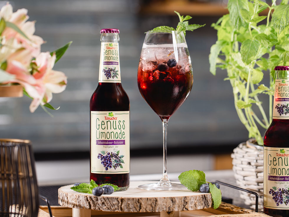 Flasche Teinacher Genuss Limonade Johannisbeere-Holunder neben einem Glas mit Eis, Beeren und Minze auf einer Holzscheibe, umgeben von Pflanzen