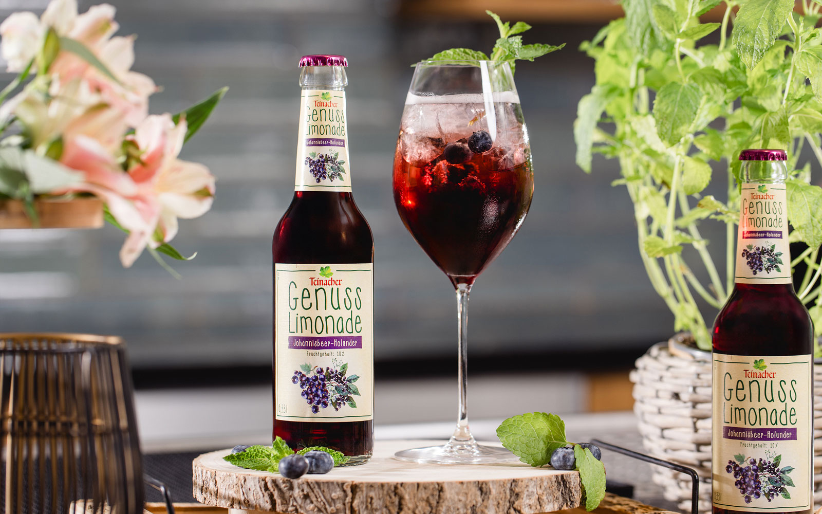 Flasche Teinacher Genuss Limonade Johannisbeere-Holunder neben einem Glas mit Eis, Beeren und Minze auf einer Holzscheibe, umgeben von Pflanzen