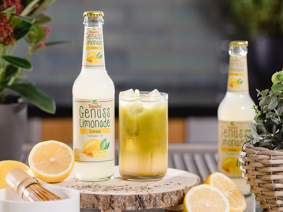 Flasche Teinacher Genuss Limonade Zitrone neben einem Glas mit Eis auf einer Holzscheibe, umgeben von Zitronen und Kräutern