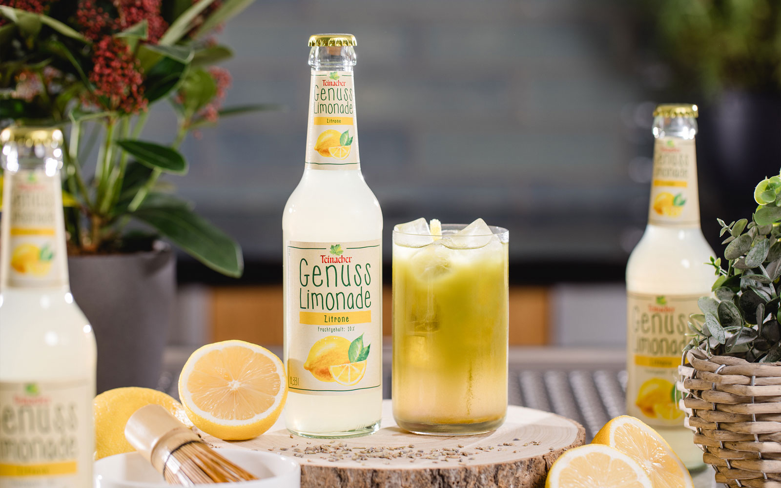 Flasche Teinacher Genuss Limonade Zitrone neben einem Glas mit Eis auf einer Holzscheibe, umgeben von Zitronen und Kräutern
