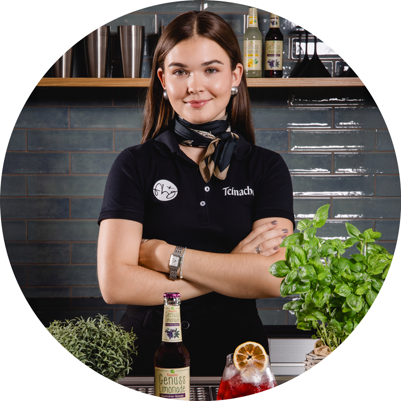 Frau mit verschränkten Armen steht hinter einer Theke mit Teinacher Getränkeflasche, Glas mit Eis und Kräutern, im Hintergrund Regal und Fliesenwand