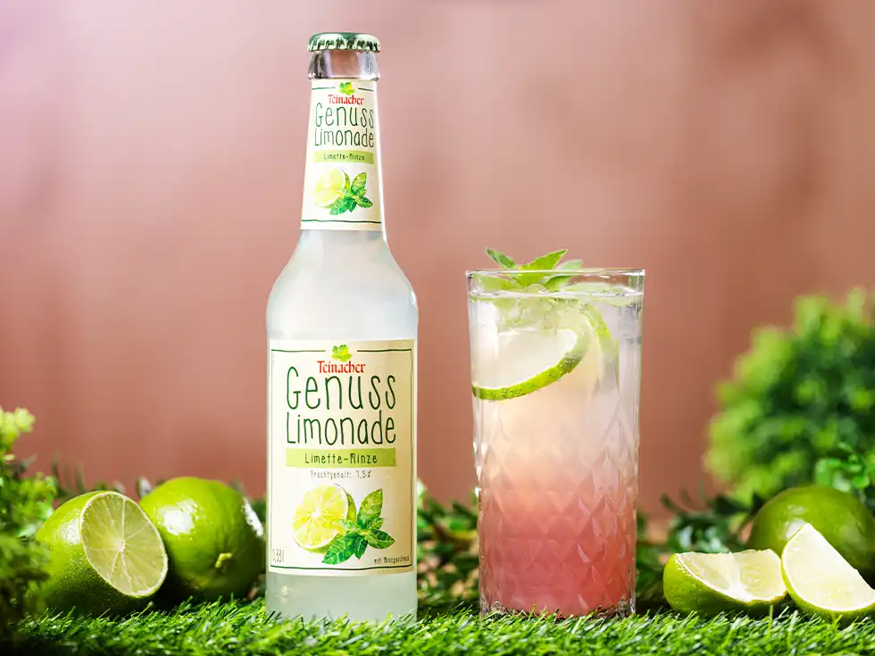 Teinacher Genusslimonade Limette-Minze neben einem Glas Mocktail mit Limettenscheiben und Minze auf grünem Untergrund.