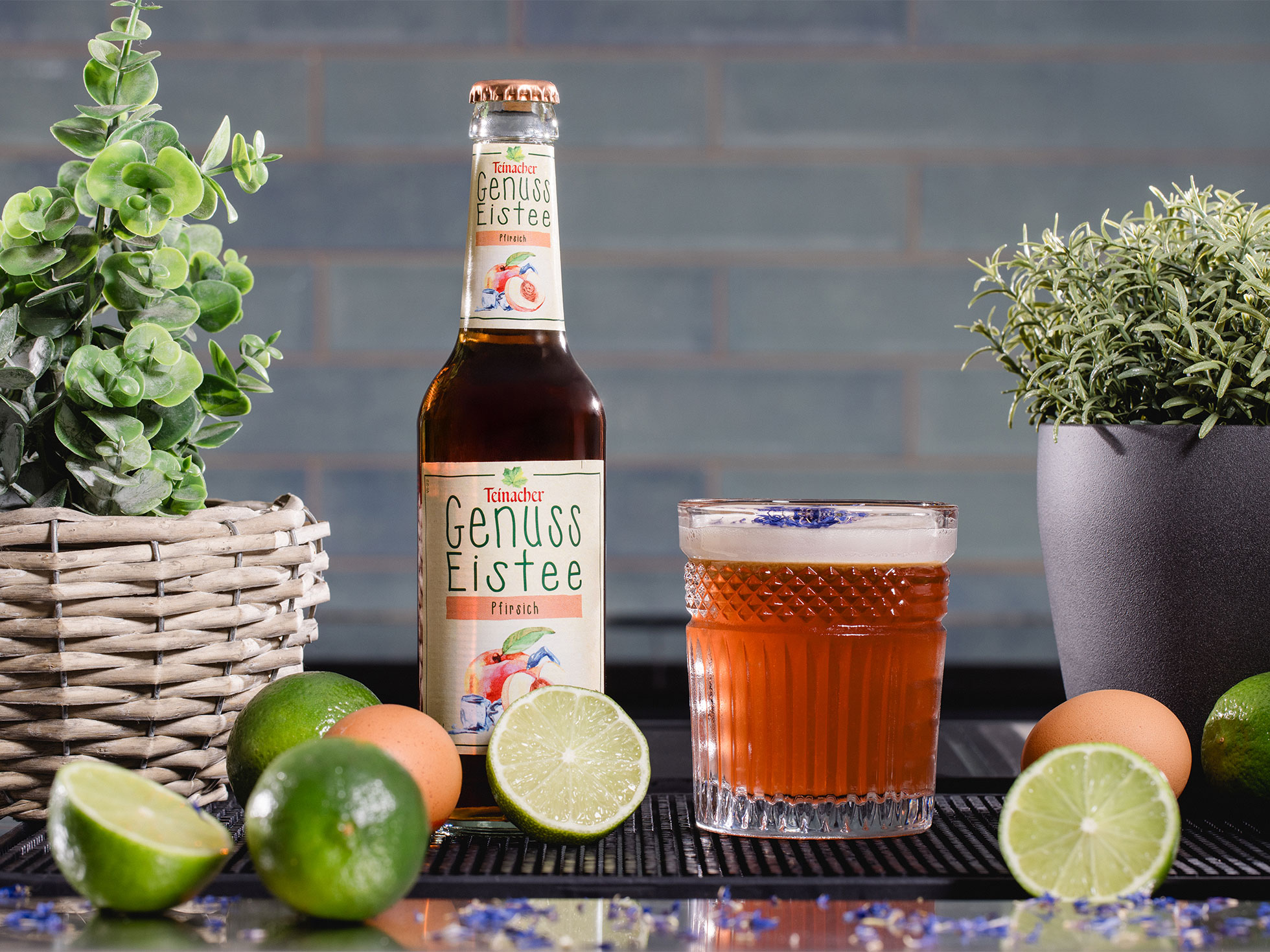 Flasche Teinacher Genuss Eistee Pfirsich neben einem gefüllten Glas, umgeben von Limetten, Eiern und Kräutern auf einer Theke
