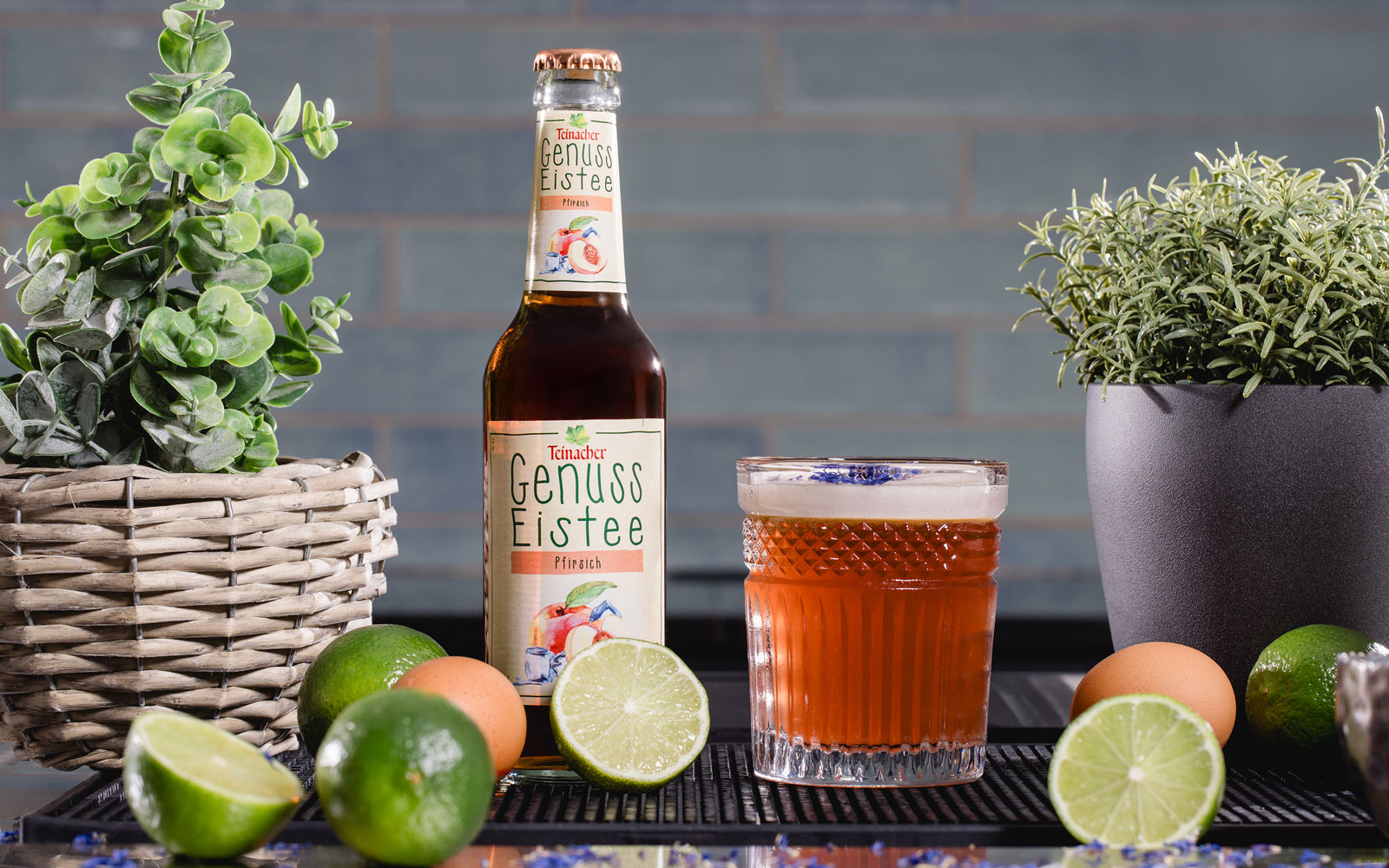 Flasche Teinacher Genuss Eistee Pfirsich neben einem gefüllten Glas, umgeben von Limetten, Eiern und Kräutern auf einer Theke