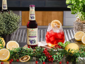 Flasche Teinacher Genuss Limonade Johannisbeere-Holunder neben einem roten Getränk mit Eis, umgeben von Zitronen, Beeren und Kräutern