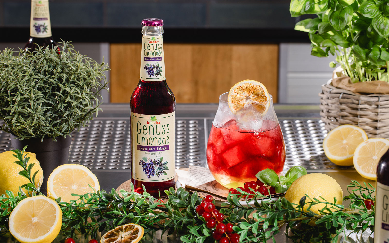 Flasche Teinacher Genuss Limonade Johannisbeere-Holunder neben einem roten Getränk mit Eis, umgeben von Zitronen, Beeren und Kräutern