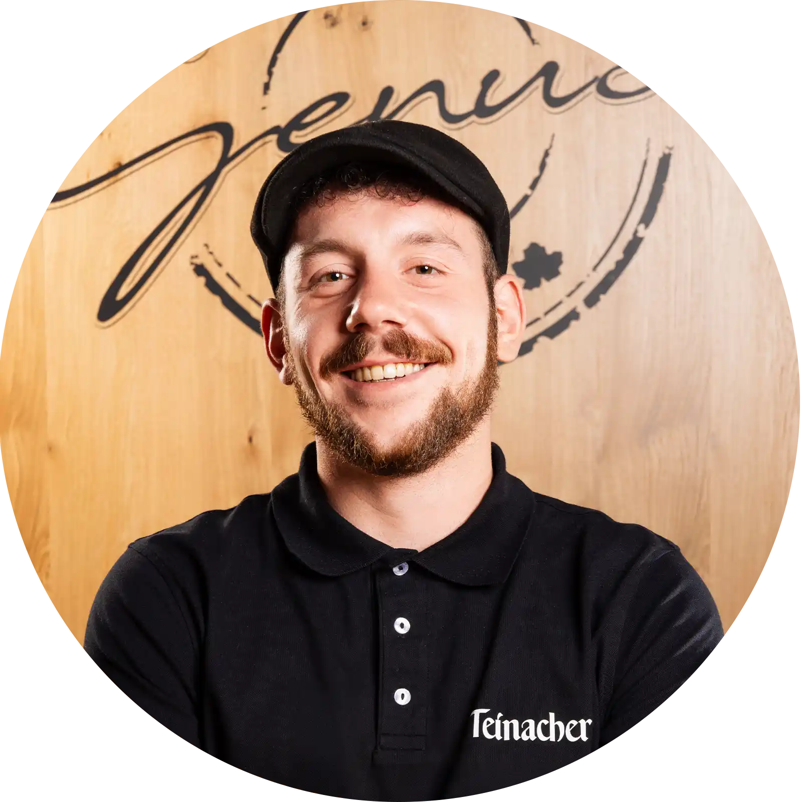 teinacher-rezepte-spritz-portrait-tobias-sutter Mann mit schwarzem Teinacher-Shirt und Mütze lächelt vor Holz-Hintergrund mit „Team Genuss“-Schriftzug.