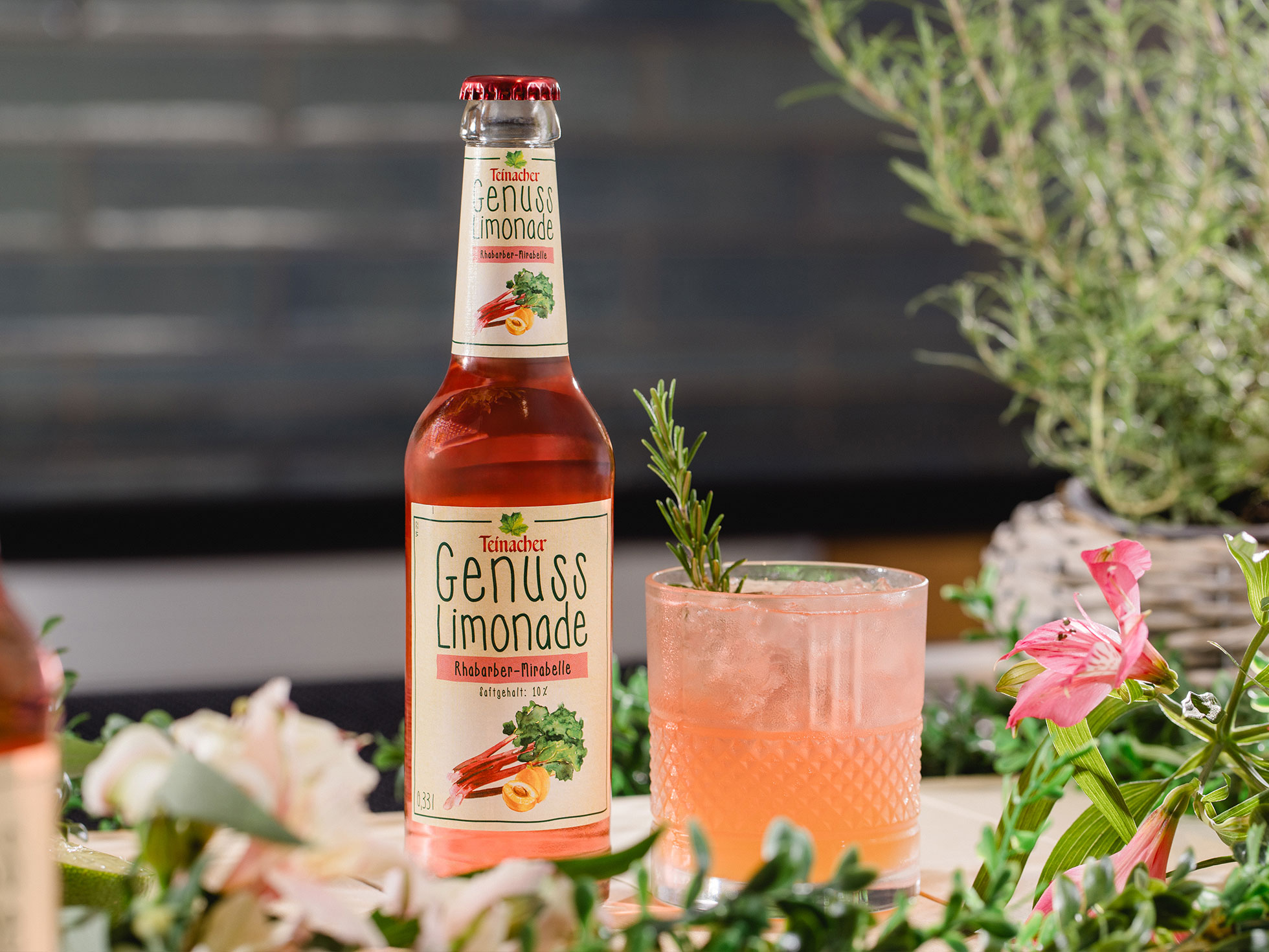 Flasche Teinacher Genuss Limonade Rhabarber-Mirabelle neben einem rosafarbenen Getränk mit Eis und Rosmarin, umgeben von Blumen und Kräutern