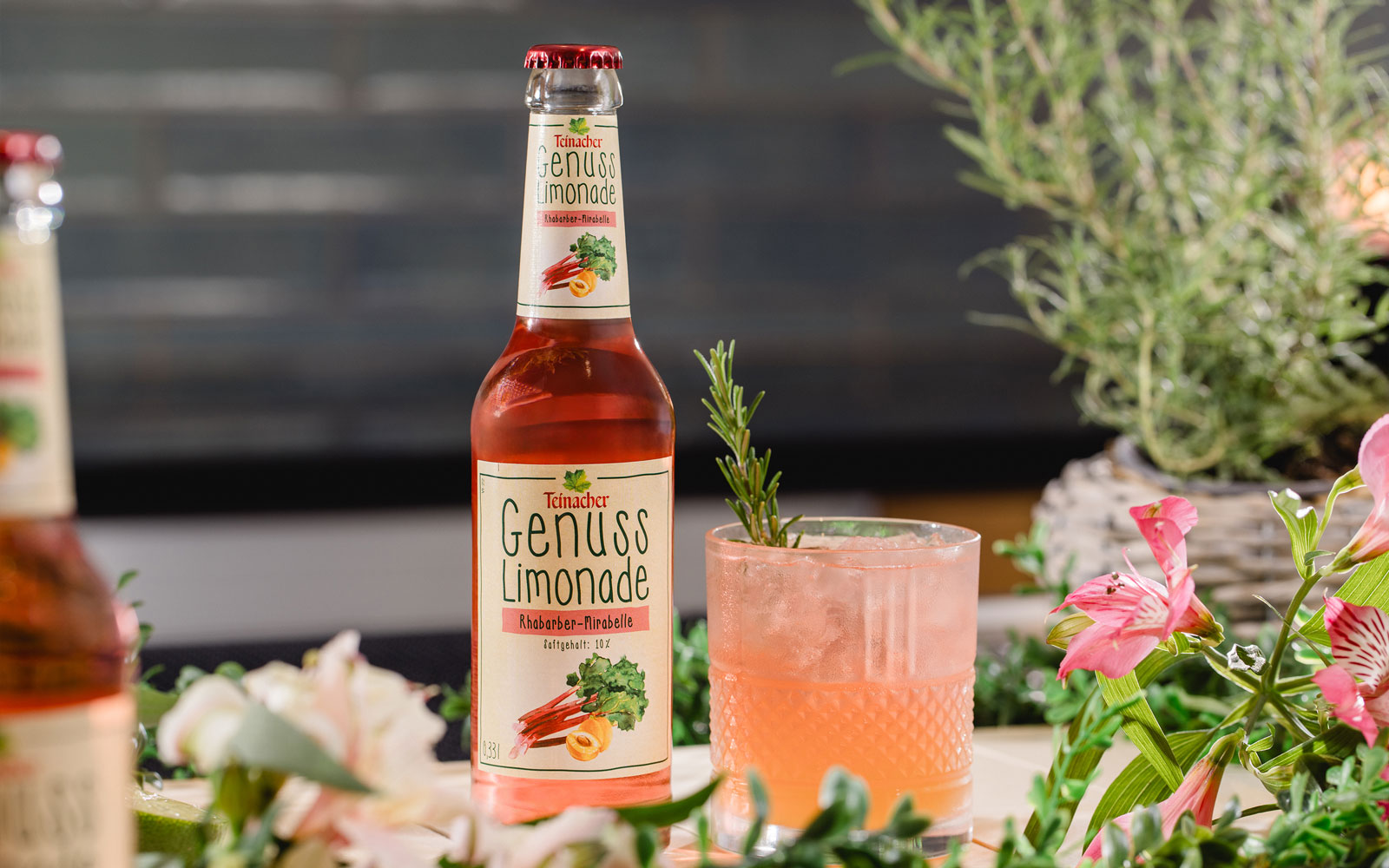 Flasche Teinacher Genuss Limonade Rhabarber-Mirabelle neben einem rosafarbenen Getränk mit Eis und Rosmarin, umgeben von Blumen und Kräutern