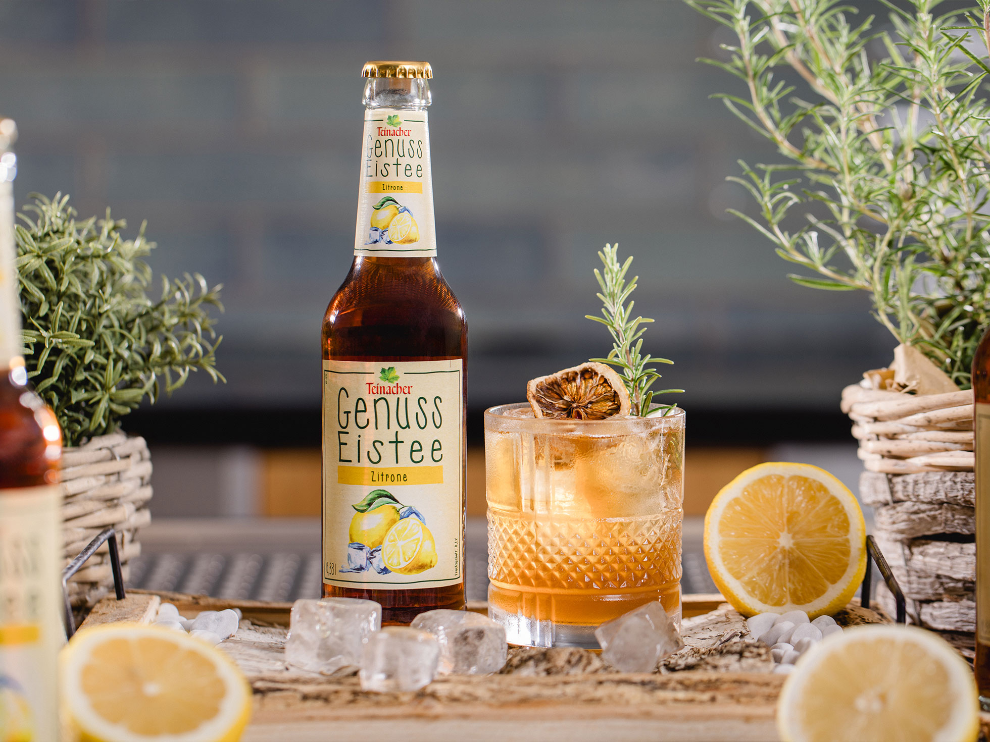 Flasche Teinacher Genuss Eistee Zitrone neben einem Glas mit Eis, Zitronenscheibe und Rosmarin, umgeben von Zitronen und Kräutern