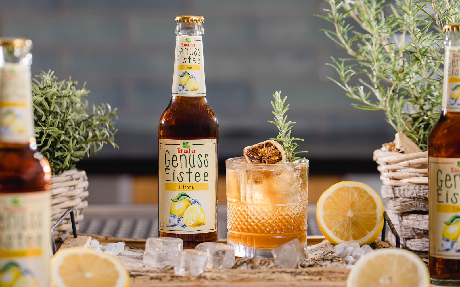 Flasche Teinacher Genuss Eistee Zitrone neben einem Glas mit Eis, Zitronenscheibe und Rosmarin, umgeben von Zitronen und Kräutern