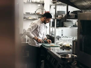 Chefkoch schneidet Gemüse in professioneller Restaurantküche vor offenem Regal mit Geschirr.