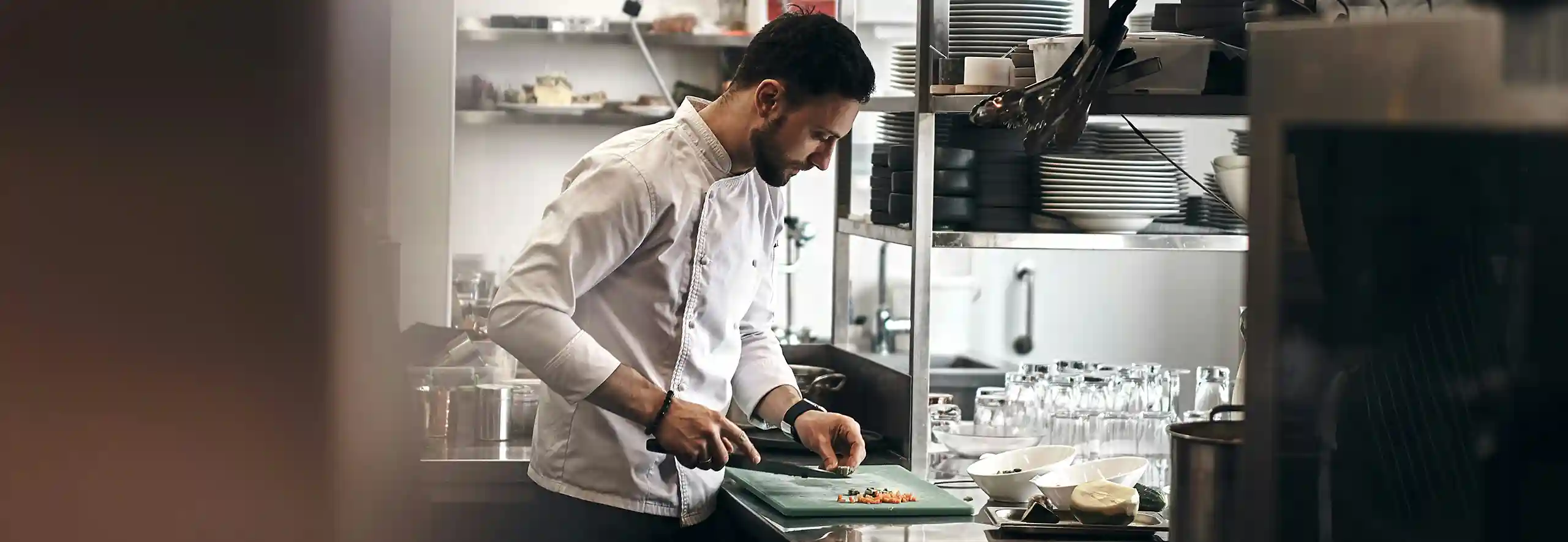 Chefkoch schneidet Gemüse in professioneller Restaurantküche vor offenem Regal mit Geschirr.