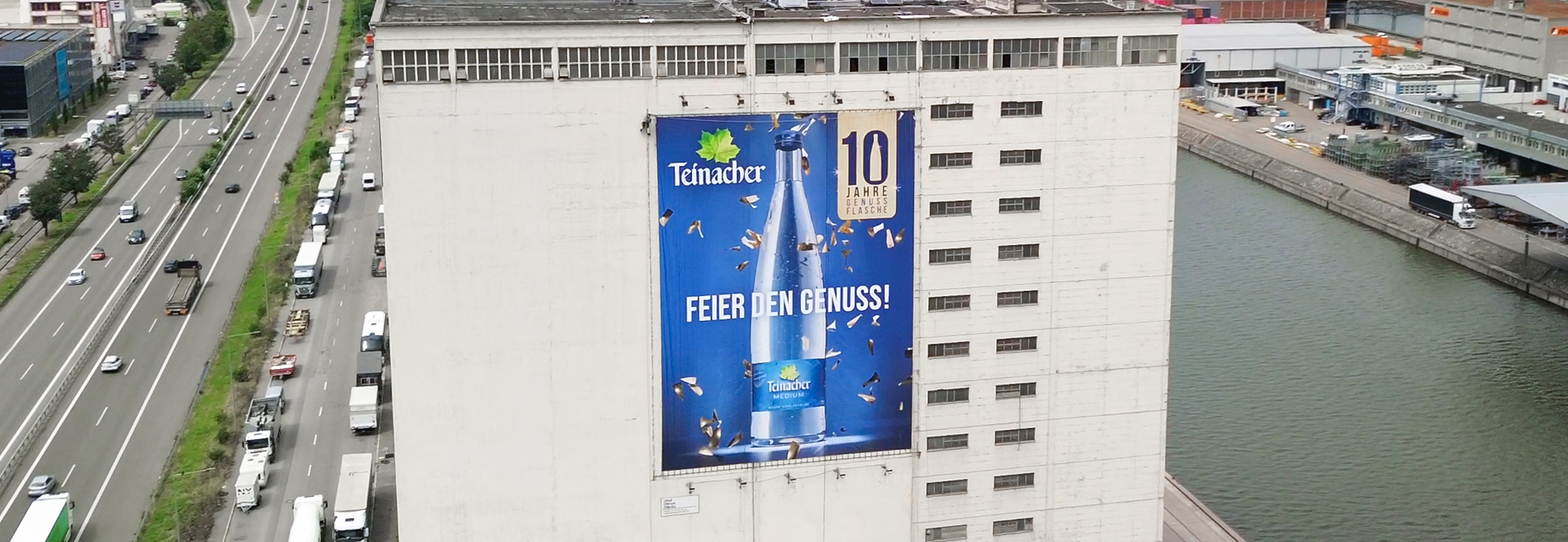 Großes Teinacher-Werbeplakat an einer Industriegebäudefassade am Fluss, mit Mineralwasserflasche und dem Motiv „10 Jahre Genussflasche“