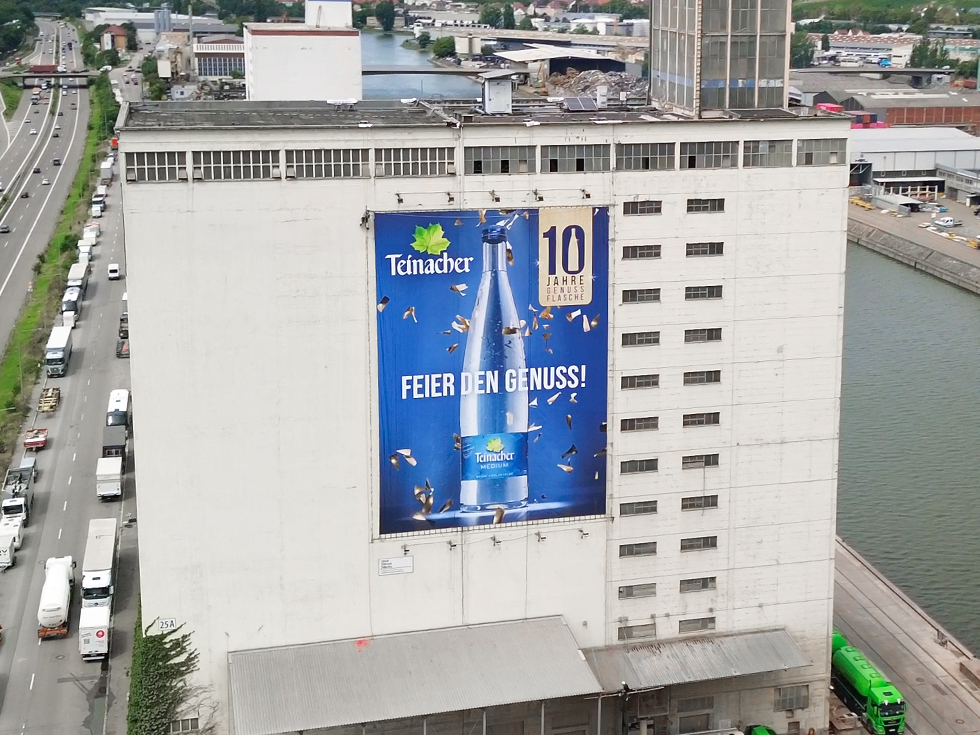 Großes Teinacher-Werbeplakat an einer Industriegebäudefassade am Fluss, mit Mineralwasserflasche und dem Motiv „10 Jahre Genussflasche“