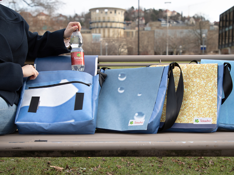 Upcycling-Taschen aus recyceltem Teinacher Werbeplakat auf einer Bank, daneben eine Teinacher Mineralwasserflasche im urbanen Umfeld.