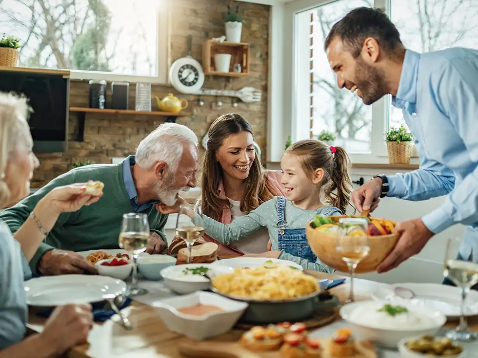 Fröhliche Familie genießt gemeinsames Essen am Tisch, Großeltern, Eltern und Kinder lachen zusammen in gemütlicher Atmosphäre