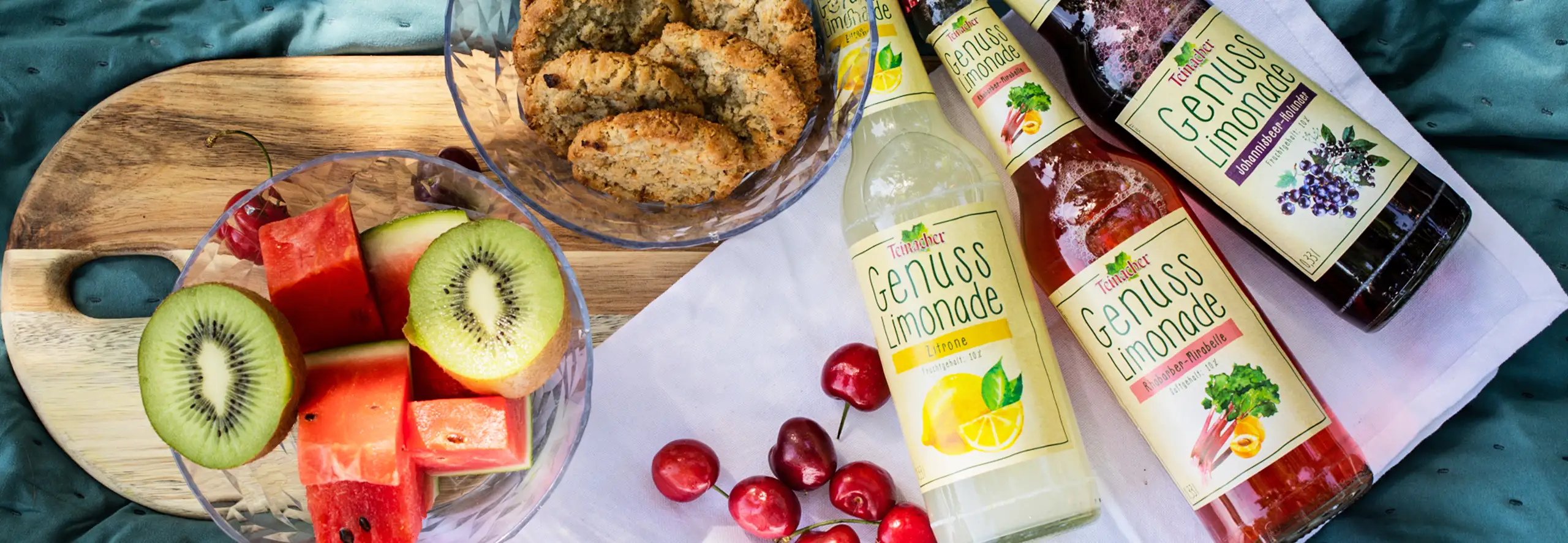 Kekse, verschiedene Früchte und drei Teinacher Genuss-Limonaden Longneckflasche 0,33 L in den Sorten Zitrone, Rhabarber-Mirabelle und Johannisbeer-Holunder.