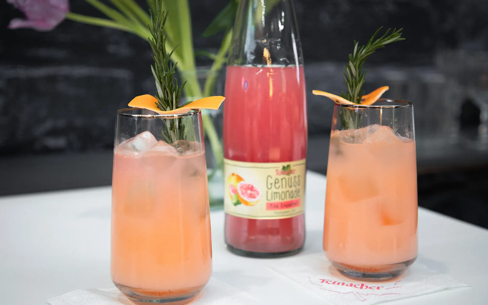 Vorderansicht einer Teinacher 0,75 Liter Pink-Grapefruit Genussflasche und 2 Gläser und Früchte im Vordergrund mit einem Blumenstrauß im Hintergrund