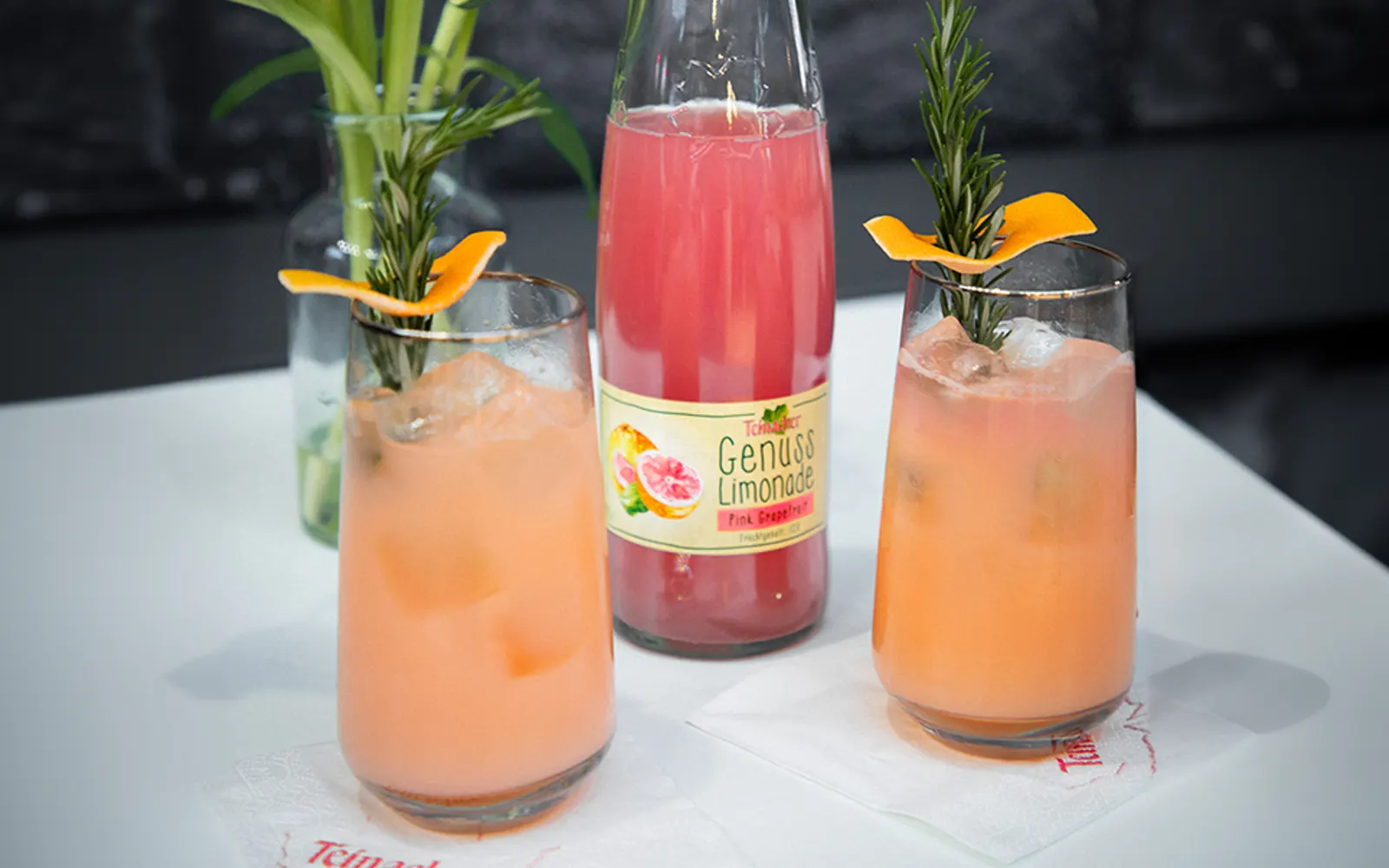 Vorderansicht einer Teinacher 0,75 Liter Pink-Grapefruit Genussflasche und 2 Gläser im Vordergrund mit einem Blumenstrauß im Hintergrund